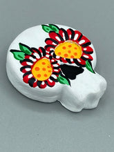 Load image into Gallery viewer, Heal emotionally and spiritually through art by honoring a loved one with a unique and beautiful Purple Spirit Piece Memorial Skull Art. Day Of The Dead Spirit Piece is an ideal addition to your ofrenda or altar that will provide a lasting tribute. These handcrafted polymer clay skull figurines are adorned with hand-painted red floral designs for the eyes. H 1 1/2” x W 1 1/2” x D 3/4”