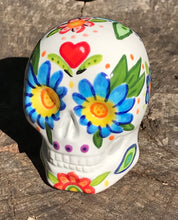 Load image into Gallery viewer, Inspired by nature, this ceramic skull statuette is full of vibrant color and intricate details. The primary color is white with Multi-color floral motifs that are painted across the skull, along with hearts and leaves and lively colorful accents around the entire skull.