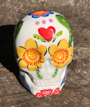 Load image into Gallery viewer, Inspired by nature, this ceramic skull statuette is full of vibrant color and intricate details. The primary color is white with multi-color floral motifs that are painted across the skull, along with hearts and leaves and lively colorful accents around the entire skull.