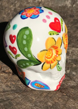 Load image into Gallery viewer, Inspired by nature, this ceramic skull statuette is full of vibrant color and intricate details. The primary color is white with multi-color floral motifs that are painted across the skull, along with hearts and leaves and lively colorful accents around the entire skull.
