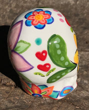 Load image into Gallery viewer, Inspired by nature, this ceramic skull statuette is full of vibrant color and intricate details. The primary color is white with multi-color floral motifs that are painted across the skull, along with hearts and leaves and lively colorful accents around the entire skull.