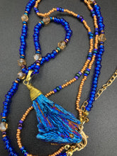 Load image into Gallery viewer, A bold deep blue kyanite wired stone pendant is surrounded by two strands of glass bead necklaces featuring a deep blue and gold shimmer palette. The shorter strand is designed to be worn as a collar or choker, while the longer second strand creates a striking contrast. With its bold appearance and eye-catching gemstone pendant, this necklace is a must-have for any jewelry collection. Necklace is 15” in length, can be adjusted to 18"