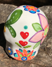 Load image into Gallery viewer, Inspired by nature, this ceramic skull statuette is full of vibrant color and intricate details. The primary color is white with multi-color floral motifs that are painted across the skull, along with hearts and leaves and lively colorful accents around the entire skull.