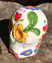 Load image into Gallery viewer, Inspired by nature, this ceramic skull statuette is full of vibrant color and intricate details. The primary color is white with multi-color floral motifs that are painted across the skull, along with hearts and leaves and lively colorful accents around the entire skull.
