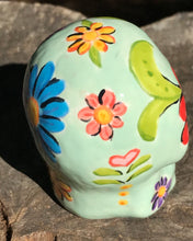 Load image into Gallery viewer, Inspired by nature, this ceramic skull statuette is full of vibrant color and intricate details. The primary color is soft pastel green with multi-color floral motifs that are painted across the skull, along with hearts and leaves and lively colorful accents around the entire skull.