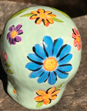 Load image into Gallery viewer, Inspired by nature, this ceramic skull statuette is full of vibrant color and intricate details. The primary color is soft pastel green with multi-color floral motifs that are painted across the skull, along with hearts and leaves and lively colorful accents around the entire skull.
