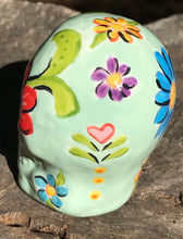Load image into Gallery viewer, Inspired by nature, this ceramic skull statuette is full of vibrant color and intricate details. The primary color is soft pastel green with multi-color floral motifs that are painted across the skull, along with hearts and leaves and lively colorful accents around the entire skull.