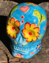 Load image into Gallery viewer, Inspired by nature, this ceramic skull statuette is full of vibrant color and intricate details. The primary color is blue with multi-color floral motifs that are painted across the skull, along with hearts and leaves and lively colorful accents around the entire skull.