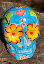 Load image into Gallery viewer, Inspired by nature, this ceramic skull statuette is full of vibrant color and intricate details. The primary color is blue with multi-color floral motifs that are painted across the skull, along with hearts and leaves and lively colorful accents around the entire skull.