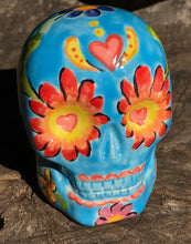 Load image into Gallery viewer, Inspired by nature, this ceramic skull statuette is full of vibrant color and intricate details. The primary color is sky blue with Multi-color floral motifs that are painted across the skull, along with hearts and leaves and lively colorful accents around the entire skull.