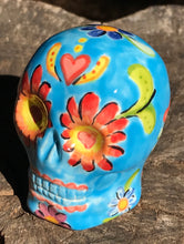 Load image into Gallery viewer, Inspired by nature, this ceramic skull statuette is full of vibrant color and intricate details. The primary color is sky blue with Multi-color floral motifs that are painted across the skull, along with hearts and leaves and lively colorful accents around the entire skull.