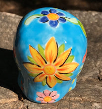 Load image into Gallery viewer, Inspired by nature, this ceramic skull statuette is full of vibrant color and intricate details. The primary color is sky blue with Multi-color floral motifs that are painted across the skull, along with hearts and leaves and lively colorful accents around the entire skull.