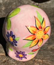 Load image into Gallery viewer, Inspired by nature, this ceramic skull statuette is full of vibrant color and intricate details. The primary color is soft pink with multi-color floral motifs that are painted across the skull, along with hearts and leaves and lively colorful accents around the entire skull.