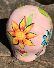Load image into Gallery viewer, Inspired by nature, this ceramic skull statuette is full of vibrant color and intricate details. The primary color is soft pink with multi-color floral motifs that are painted across the skull, along with hearts and leaves and lively colorful accents around the entire skull.