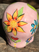 Load image into Gallery viewer, Inspired by nature, this ceramic skull statuette is full of vibrant color and intricate details. The primary color is soft pink with multi-color floral motifs that are painted across the skull, along with hearts and leaves and lively colorful accents around the entire skull.