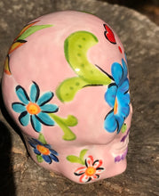 Load image into Gallery viewer, Inspired by nature, this ceramic skull statuette is full of vibrant color and intricate details. The primary color is soft pink with multi-color floral motifs that are painted across the skull, along with hearts and leaves and lively colorful accents around the entire skull.
