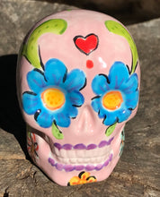 Load image into Gallery viewer, Inspired by nature, this ceramic skull statuette is full of vibrant color and intricate details. The primary color is soft pink with multi-color floral motifs that are painted across the skull, along with hearts and leaves and lively colorful accents around the entire skull.