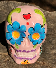 Load image into Gallery viewer, Inspired by nature, this ceramic skull statuette is full of vibrant color and intricate details. The primary color is soft pink with multi-color floral motifs that are painted across the skull, along with hearts and leaves and lively colorful accents around the entire skull.