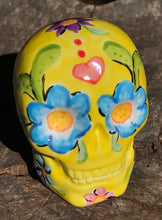 Load image into Gallery viewer, Inspired by nature, this ceramic skull statuette is full of vibrant color and intricate details. The primary color is yellow with Multi-color floral motifs that are painted across the skull, along with hearts and leaves and lively colorful accents around the entire skull.