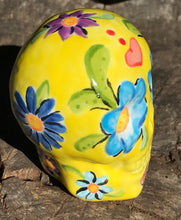 Load image into Gallery viewer, Inspired by nature, this ceramic skull statuette is full of vibrant color and intricate details. The primary color is yellow with Multi-color floral motifs that are painted across the skull, along with hearts and leaves and lively colorful accents around the entire skull.