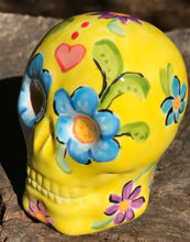 Load image into Gallery viewer, Inspired by nature, this ceramic skull statuette is full of vibrant color and intricate details. The primary color is yellow with Multi-color floral motifs that are painted across the skull, along with hearts and leaves and lively colorful accents around the entire skull.