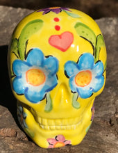 Load image into Gallery viewer, Inspired by nature, this ceramic skull statuette is full of vibrant color and intricate details. The primary color is yellow with Multi-color floral motifs that are painted across the skull, along with hearts and leaves and lively colorful accents around the entire skull.