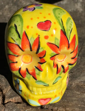 Load image into Gallery viewer, This ceramic skull statuette is a stunning piece of art inspired by nature. Its vibrant yellow hue is decorated with multi-color floral motifs, hearts, leaves and colorful accents, creating a beautiful piece of hand-painted, Day of the Dead-themed art. Measurements: H 2 3/4” x W 2” x D 2”