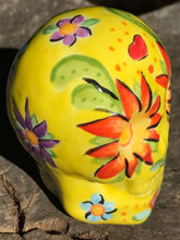 Load image into Gallery viewer, This ceramic skull statuette is a stunning piece of art inspired by nature. Its vibrant yellow hue is decorated with multi-color floral motifs, hearts, leaves and colorful accents, creating a beautiful piece of hand-painted, Day of the Dead-themed art. Measurements: H 2 3/4” x W 2” x D 2”