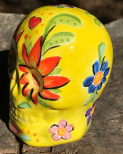 Load image into Gallery viewer, This ceramic skull statuette is a stunning piece of art inspired by nature. Its vibrant yellow hue is decorated with multi-color floral motifs, hearts, leaves and colorful accents, creating a beautiful piece of hand-painted, Day of the Dead-themed art. Measurements: H 2 3/4” x W 2” x D 2”