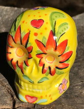 Load image into Gallery viewer, This ceramic skull statuette is a stunning piece of art inspired by nature. Its vibrant yellow hue is decorated with multi-color floral motifs, hearts, leaves and colorful accents, creating a beautiful piece of hand-painted, Day of the Dead-themed art. Measurements: H 2 3/4” x W 2” x D 2”