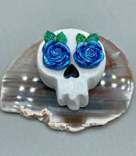 Load image into Gallery viewer, Cremation memorial art, such as Day of the Dead Spirit Pieces, provide a unique way to keep a loved one close to your heart. Handcrafted with care, each piece of artwork is specially tailored to the individual, providing a lasting reminder of those who are dearly missed. On altars and ofrendas, these pieces make a beautiful addition. Composed with meticulous attention to detail, these mini polymer clay skulls feature hand sculpted bright blue roses and leaves. H 1 1/2” x W 1 1/2” x D 3/4”