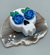Load image into Gallery viewer, Cremation memorial art, such as Day of the Dead Spirit Pieces, provide a unique way to keep a loved one close to your heart. Handcrafted with care, each piece of artwork is specially tailored to the individual, providing a lasting reminder of those who are dearly missed. On altars and ofrendas, these pieces make a beautiful addition. Composed with meticulous attention to detail, these mini polymer clay skulls feature hand sculpted bright blue roses and leaves. H 1 1/2” x W 1 1/2” x D 3/4”