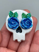 Load image into Gallery viewer, Cremation memorial art, such as Day of the Dead Spirit Pieces, provide a unique way to keep a loved one close to your heart. Handcrafted with care, each piece of artwork is specially tailored to the individual, providing a lasting reminder of those who are dearly missed. On altars and ofrendas, these pieces make a beautiful addition. Composed with meticulous attention to detail, these mini polymer clay skulls feature hand sculpted bright blue roses and leaves. H 1 1/2” x W 1 1/2” x D 3/4”