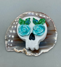 Load image into Gallery viewer, Cremation memorial art, such as Day of the Dead Spirit Pieces, provide a unique way to keep a loved one close to your heart. Handcrafted with care, each piece of artwork is specially tailored to the individual, providing a lasting reminder of those who are dearly missed. On altars and ofrendas, these pieces make a beautiful addition. Composed with meticulous attention to detail, these mini polymer clay skulls feature hand sculpted light blue roses and leaves. H 1 1/2” x W 1 1/2” x D 3/4”