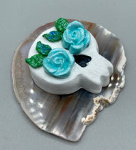 Load image into Gallery viewer, Cremation memorial art, such as Day of the Dead Spirit Pieces, provide a unique way to keep a loved one close to your heart. Handcrafted with care, each piece of artwork is specially tailored to the individual, providing a lasting reminder of those who are dearly missed. On altars and ofrendas, these pieces make a beautiful addition. Composed with meticulous attention to detail, these mini polymer clay skulls feature hand sculpted light blue roses and leaves. H 1 1/2” x W 1 1/2” x D 3/4”