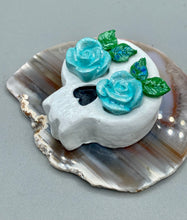 Load image into Gallery viewer, Cremation memorial art, such as Day of the Dead Spirit Pieces, provide a unique way to keep a loved one close to your heart. Handcrafted with care, each piece of artwork is specially tailored to the individual, providing a lasting reminder of those who are dearly missed. On altars and ofrendas, these pieces make a beautiful addition. Composed with meticulous attention to detail, these mini polymer clay skulls feature hand sculpted light blue roses and leaves. H 1 1/2” x W 1 1/2” x D 3/4”