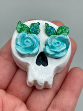 Load image into Gallery viewer, Cremation memorial art, such as Day of the Dead Spirit Pieces, provide a unique way to keep a loved one close to your heart. Handcrafted with care, each piece of artwork is specially tailored to the individual, providing a lasting reminder of those who are dearly missed. On altars and ofrendas, these pieces make a beautiful addition. Composed with meticulous attention to detail, these mini polymer clay skulls feature hand sculpted light blue roses and leaves. H 1 1/2” x W 1 1/2” x D 3/4”