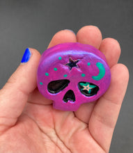 Load image into Gallery viewer, Cute and spooky magnet design to adorn your refrigerator, lockers, workstation, toolbox, or dishwasher. This skull statuette is a tribute to nature. Its metallic violet hue evokes sundown, transitioning the sky from blue to purple to black. Adding to the appeal of this piece is a teal-green moon and stars that provide a dramatic contrast against the violet. The forehead is finished with a rainbow hematite star bead for a unique accent.  H 1 1/2” x W 1 1/2” x D 3/4”