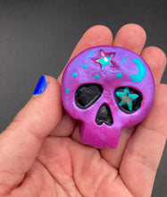 Load image into Gallery viewer, Cute and spooky magnet design to adorn your refrigerator, lockers, workstation, toolbox, or dishwasher. This skull statuette is a tribute to nature. Its metallic violet hue evokes sundown, transitioning the sky from blue to purple to black. Adding to the appeal of this piece is a teal-green moon and stars that provide a dramatic contrast against the violet. The forehead is finished with a rainbow hematite star bead for a unique accent.  H 1 1/2” x W 1 1/2” x D 3/4”
