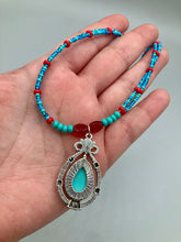 Load image into Gallery viewer, This captivating Southwestern style necklace features a two-strand design of bold and vibrant turquoise and red glass beads. At the center, a bright turquoise pendant is framed in a distinctive design. Measurements: Necklace is 16 1/2 ” adjustable up to 18"