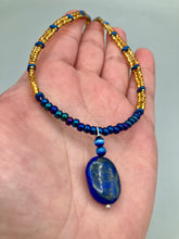 Load image into Gallery viewer, This Lapis pendant necklace features dark blue cats eye beads and two strands of gold glass beads with dark blue double hole beads. The large multi-colored glass beads provide remarkable shimmer and a full range of colors, such as purples, blues, greens, and golds, that enhances the lapis stone pendant. Measurements: Necklace is 19” in length