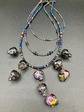 Load image into Gallery viewer, Massively beautiful and bold skull necklace is the ultimate statement piece to add to your jewelry collection! Three strands necklace, contains 5 strands of large skull beads that give a cascade look to this beautiful necklace. Edgy gun metal faceted shimmery multi-color glass hues of silver, purple, pink, blue, and gold. This bold and beautiful necklace drapes across your neck, comes with dainty matching earrings to complete the look. Necklace is 15 1/2” adjustable up to 18" - glass skulls are 5/8”
