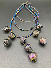 Load image into Gallery viewer, Massively beautiful and bold skull necklace is the ultimate statement piece to add to your jewelry collection! Three strands necklace, contains 5 strands of large skull beads that give a cascade look to this beautiful necklace. Edgy gun metal faceted shimmery multi-color glass hues of silver, purple, pink, blue, and gold. This bold and beautiful necklace drapes across your neck, comes with dainty matching earrings to complete the look. Necklace is 15 1/2” adjustable up to 18" - glass skulls are 5/8”