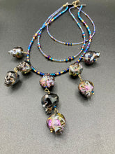 Load image into Gallery viewer, Massively beautiful and bold skull necklace is the ultimate statement piece to add to your jewelry collection! Three strands necklace, contains 5 strands of large skull beads that give a cascade look to this beautiful necklace. Edgy gun metal faceted shimmery multi-color glass hues of silver, purple, pink, blue, and gold. This bold and beautiful necklace drapes across your neck, comes with dainty matching earrings to complete the look. Necklace is 15 1/2” adjustable up to 18" - glass skulls are 5/8”