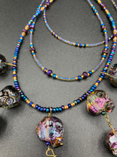 Load image into Gallery viewer, Massively beautiful and bold skull necklace is the ultimate statement piece to add to your jewelry collection! Three strands necklace, contains 5 strands of large skull beads that give a cascade look to this beautiful necklace. Edgy gun metal faceted shimmery multi-color glass hues of silver, purple, pink, blue, and gold. This bold and beautiful necklace drapes across your neck, comes with dainty matching earrings to complete the look. Necklace is 15 1/2” adjustable up to 18" - glass skulls are 5/8”