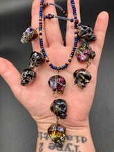 Load image into Gallery viewer, Massively beautiful and bold skull necklace is the ultimate statement piece to add to your jewelry collection! Three strands necklace, contains 5 strands of large skull beads that give a cascade look to this beautiful necklace. Edgy gun metal faceted shimmery multi-color glass hues of silver, purple, pink, blue, and gold. This bold and beautiful necklace drapes across your neck, comes with dainty matching earrings to complete the look. Necklace is 15 1/2” adjustable up to 18" - glass skulls are 5/8”