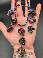 Load image into Gallery viewer, Massively beautiful and bold skull necklace is the ultimate statement piece to add to your jewelry collection! Three strands necklace, contains 5 strands of large skull beads that give a cascade look to this beautiful necklace. Edgy gun metal faceted shimmery multi-color glass hues of silver, purple, pink, blue, and gold. This bold and beautiful necklace drapes across your neck, comes with dainty matching earrings to complete the look. Necklace is 15 1/2” adjustable up to 18" - glass skulls are 5/8”