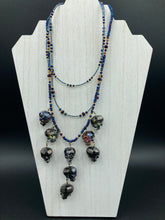 Load image into Gallery viewer, Massively beautiful and bold skull necklace is the ultimate statement piece to add to your jewelry collection! Three strands necklace, contains 5 strands of large skull beads that give a cascade look to this beautiful necklace. Edgy gun metal faceted shimmery multi-color glass hues of silver, purple, pink, blue, and gold. This bold and beautiful necklace drapes across your neck, comes with dainty matching earrings to complete the look. Necklace is 15 1/2” adjustable up to 18" - glass skulls are 5/8”