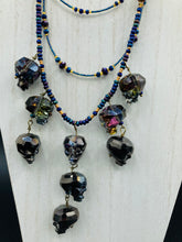 Load image into Gallery viewer, Massively beautiful and bold skull necklace is the ultimate statement piece to add to your jewelry collection! Three strands necklace, contains 5 strands of large skull beads that give a cascade look to this beautiful necklace. Edgy gun metal faceted shimmery multi-color glass hues of silver, purple, pink, blue, and gold. This bold and beautiful necklace drapes across your neck, comes with dainty matching earrings to complete the look. Necklace is 15 1/2” adjustable up to 18" - glass skulls are 5/8”