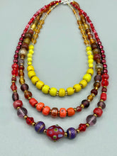 Load image into Gallery viewer, This necklace consists of three strands with a multitude of shades ranging from red to orange, gold, and amber, providing a captivating visual combination. Crafted with a combination of glass, ceramic and metal beads, this eye-catching piece of jewelry radiates power and strength. Measurements: Necklace is 16” in length, can be adjusted up to 17 1/2"